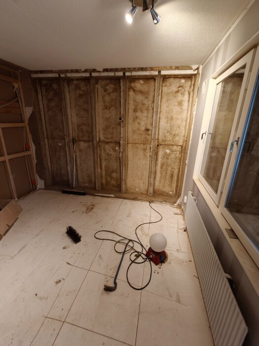 Rum under renovering med demolering av vägg, synliga reglar och verktyg på golvet, inklusive borste och arbetslampa.