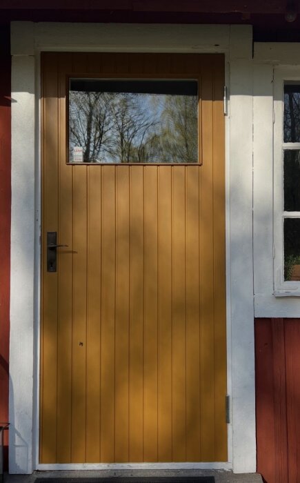Gul dörr med stående smal träpanel och litet fönster, placerad i en röd och vit fasad, trä omger hela dörren utan sparkplåt.