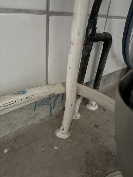 Vita rör och ett svart, äldre rör syns intill kaklade väggar och betonggolv, färg och smutsdetaljer på vägg och rör.