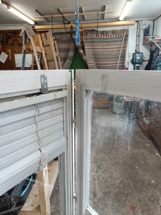 Gammalt fönster halvöppet i en verkstad med smutsiga rutor och trasig persienn, omgiven av verktyg och material i bakgrunden.