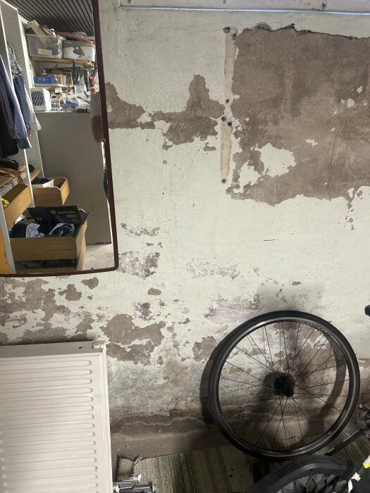 Avskalad vägg med rostfläckar, cykelhjul lutat mot väggen, och en spegel som reflekterar en del av rummet med verktyg och förvaringslådor.