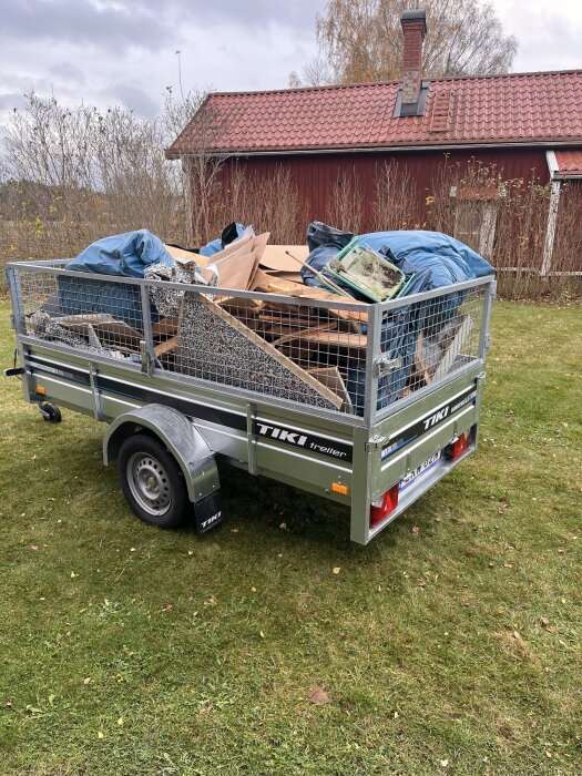 En släpvagn fylld med olika föremål, såsom täcken och byggmaterial, står parkerad på en gräsmatta framför ett rött hus.