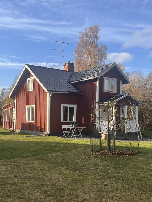 Rött trähus med vita foder och tegelsten skorsten, omgiven av grönt gräs och träd. En vit trädgårdsstol och bord i trädgården.