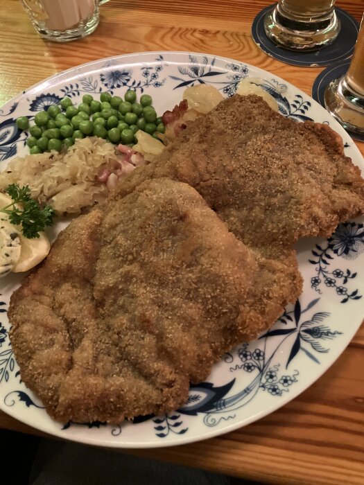 En tallrik med Wienerschnitzel, potatis, ärtor och surkål, serverad på en träbord med ölglas i bakgrunden.