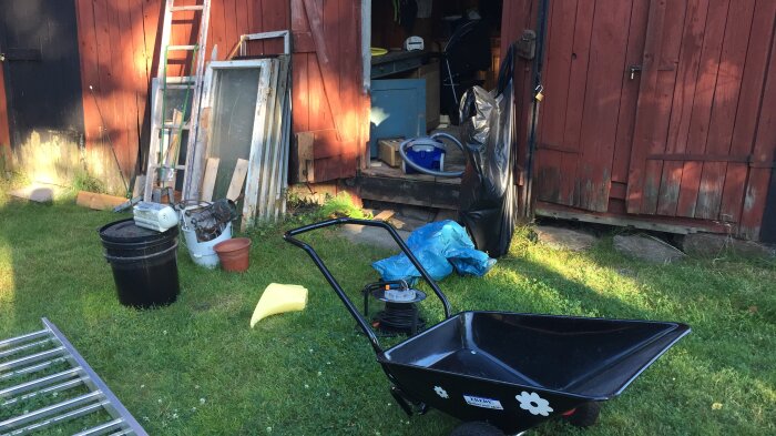 Svart skottkärra med blommor framför en röd träbyggnad. Omgiven av trädgårdsredskap, fönsterramar och trälådor.