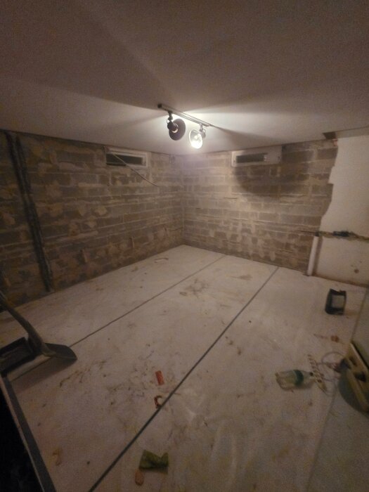 Källarutrymme under renovering med exponerade tegelväggar och oavslutat golv; takbelysning med två spotlights.