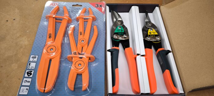Två förpackningar med byggverktyg: en med tre orange slangklämmor och en med två plåtsaxar i svart och orange.
