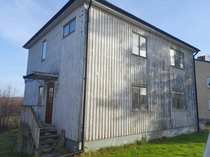 Gammalt hus med sliten stående lockpanel och smala lockbrädor, fönster utan tydliga foder och en presenning på betongtrappan.