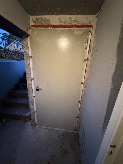 Nyinstallerad dörr i ett trångt utrymme, omgiven av omålad och ojämn vägg. Trappa synlig till vänster.
