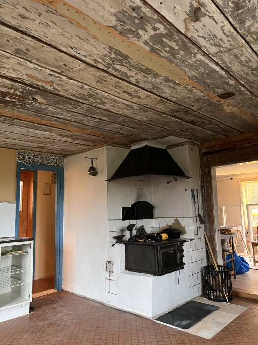 Rustikt kökstak med flagnande vit färg, synliga plankor och gammaldags vedspis i hörnet av ett kök under renovering.