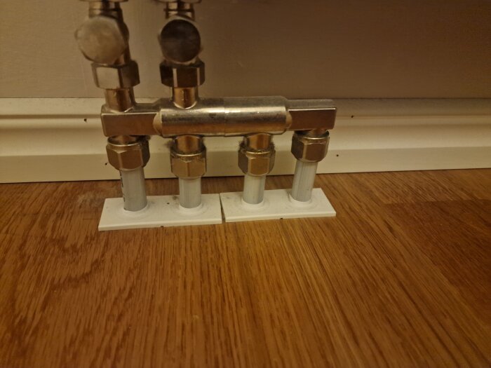 Röranslutningar under ett element med metallkopplingar och vita plaströr som kommer upp ur golvet, intill en vit vägglist på ett trägolv.