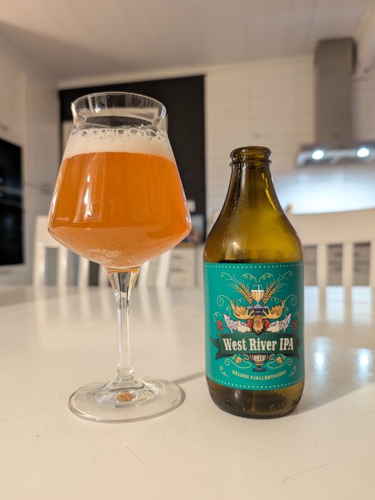 Ett glas öl och en flaska med etiketten "West River IPA" står på ett bord i ett kök.