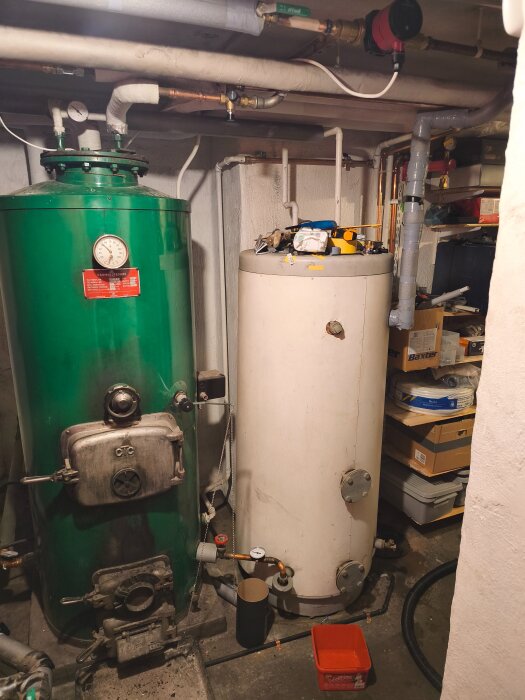 Grön och vit tank i pannrum med rörsystem, tryckmätare och cirkulationspump. Verktyg och material ovanpå den vita tanken.