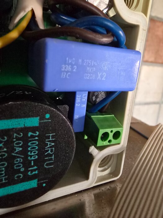 Elektrisk komponentbox med kondensator, blå och brun koppartråd samt en grön kontakt med två ingångar, märkta med text och symboler.