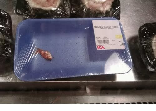 En mycket liten fisk på ett blått plasttråg från ICA, inslagen i plastfilm, ligger på en butiksdisk för fiskprodukter.