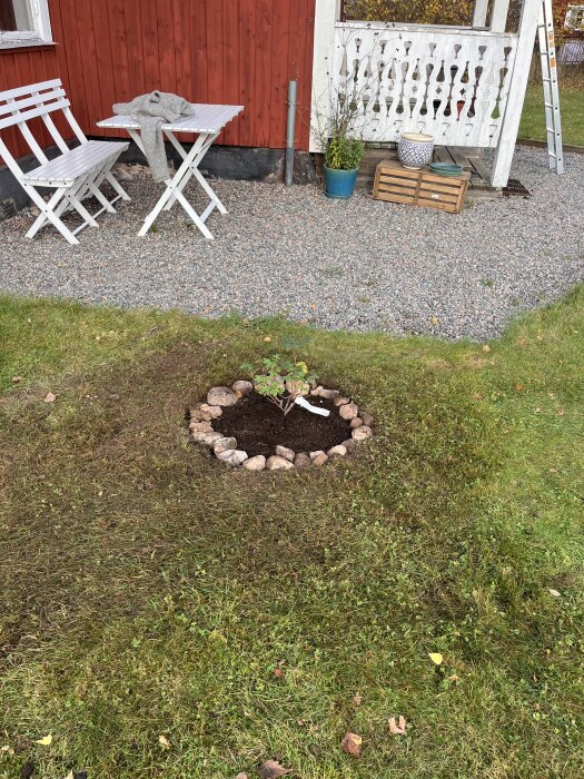Liten perennrabatt med stenkrans på gräsmatta nära röd byggnad med vita möbler och trädäck.