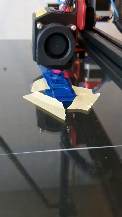 En pågående 3D-printning visas där ett blått objekt framträder. Tejp har använts för att fixera delar som lossnat under printprocessen.