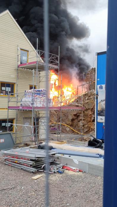 En byggnadsställning vid en gul byggnad, tjock svart rök och eldslågor syns bakom. En allvarlig brand pågår.