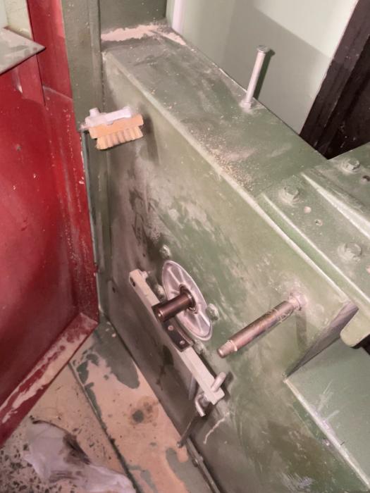 Tjock, grön metallisk dörr med säkerhetslås och spak, slitage och smuts syns. Stängd och robust, röda detaljer intill.