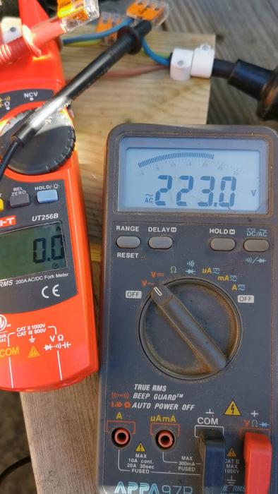 Två multimeters visar mätvärden: en ampere, den andra spänning. Elektriska anslutningar och kablar syns i bakgrunden.