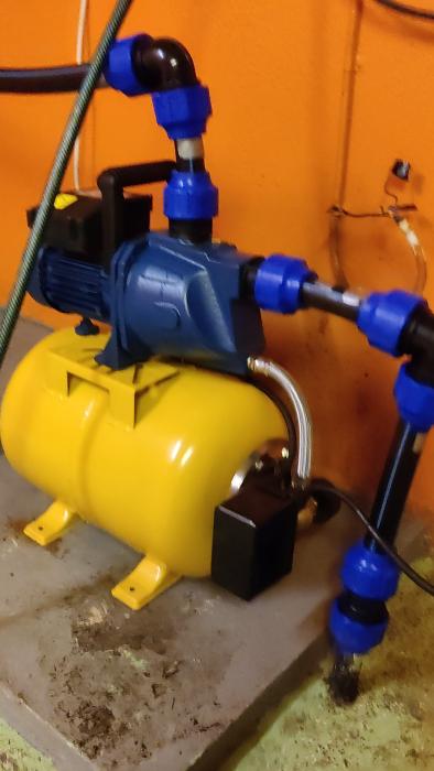 En gul luftkompressor med blå anslutningar står på en betongbotten framför en orange vägg.