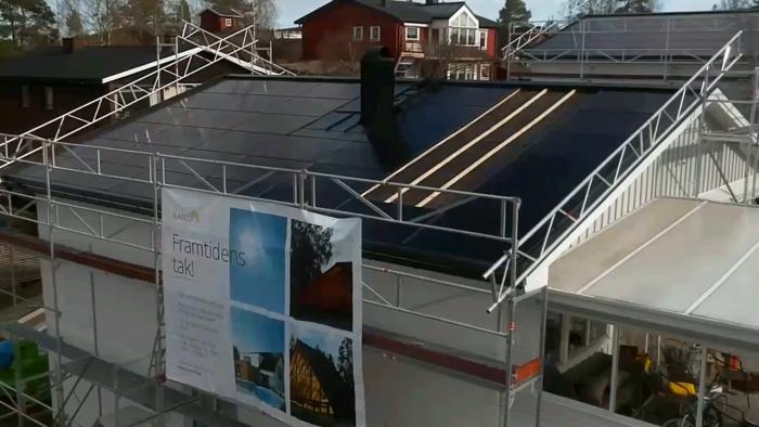 En byggnadsställning runt ett hus med solpaneler på taket. En reklambanner annonserar "Framtidens tak!"