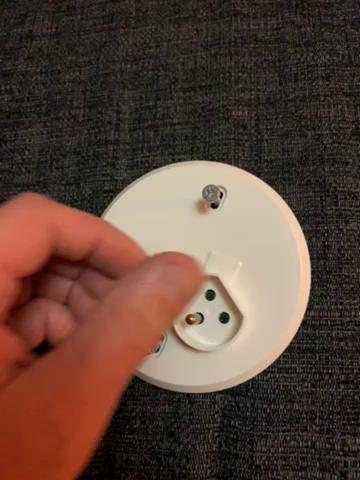 En hand håller en vit rund enhet med en Google-logotyp, troligen en Google Nest Mini, på en mörk textilerad yta.