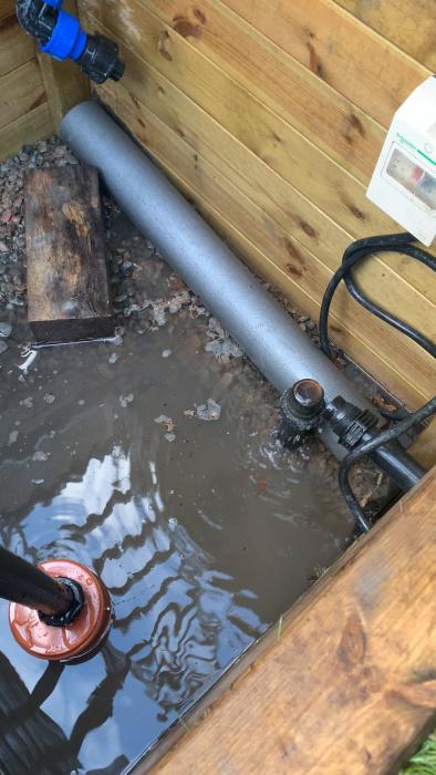 En vattenpump ansluten till en grå rörledning intill en trävägg, ovanför en vattenansamling med löv. En timer-switch syns på väggen.