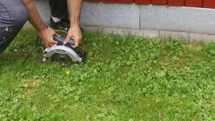 En person använder en cirkelsåg för att såga i gräset nära en stenmur.