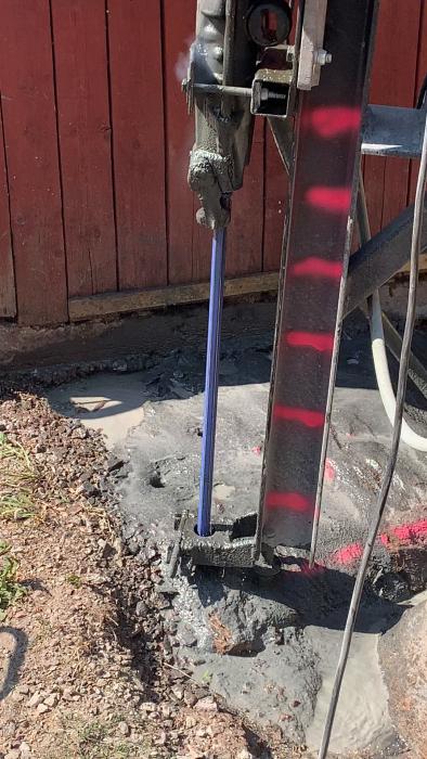 En betongpump pumpar grå betong genom en blå slang, bredvid en rödstaket, på en byggarbetsplats med synlig grus och jord.