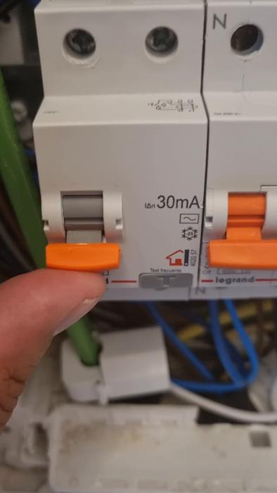 En person trycker på en orange knapp på en vit elektrisk kretsavbrytare i ett elskåp med kablar.