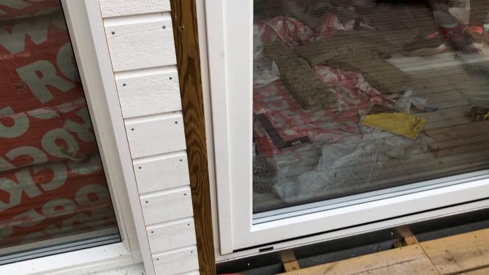 En öppen vit dörr med en reflektion av en person, leder till en veranda under konstruktion, med täckta fönster och konstruktionsmaterial synliga.