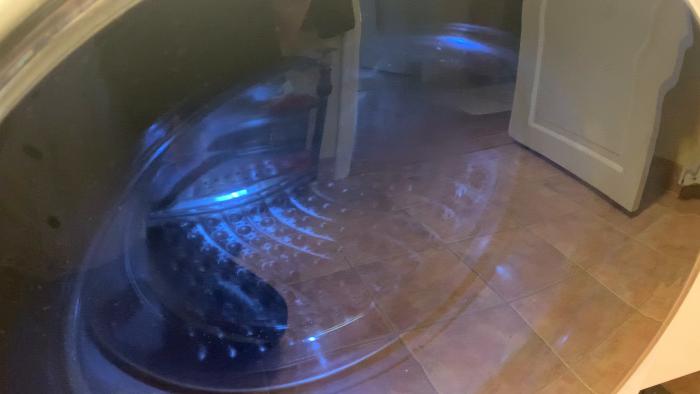 Filmen visar en tvättmaskin i rörelse, reflekterad på en blank klinkergolv, skapar ljuseffekter och suddiga reflektioner genom en glasyta.