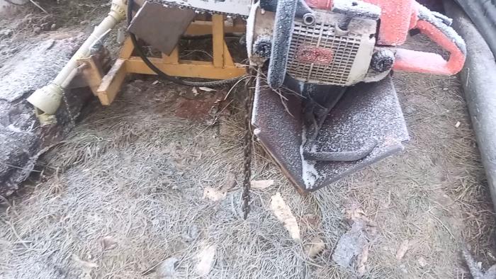 En motorsåg täckt av is och snö, vilket tyder på kallt väder, placerad utomhus nära ved och redskap.