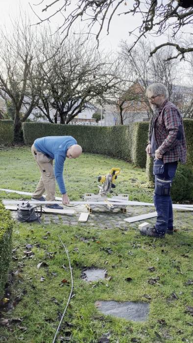 Två män arbetar utomhus, byggmaterial och verktyg på gräset, trädgårdsarbete eller renoveringsprojekt pågår.