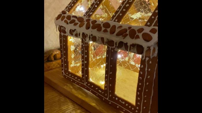 Ett pepparkakshus med lysande ljusslingor inuti, dekorativt glasyr och färgrika detaljer syns på ett mörkt underlag.