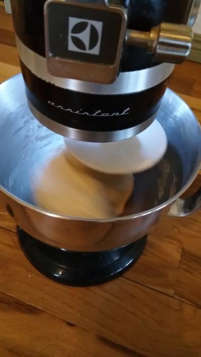 En köksmaskin blandar ingredienser i en rostfri skål på ett träköksbord. Maskinen är i funktion med en visp- eller blandningsverktyg.