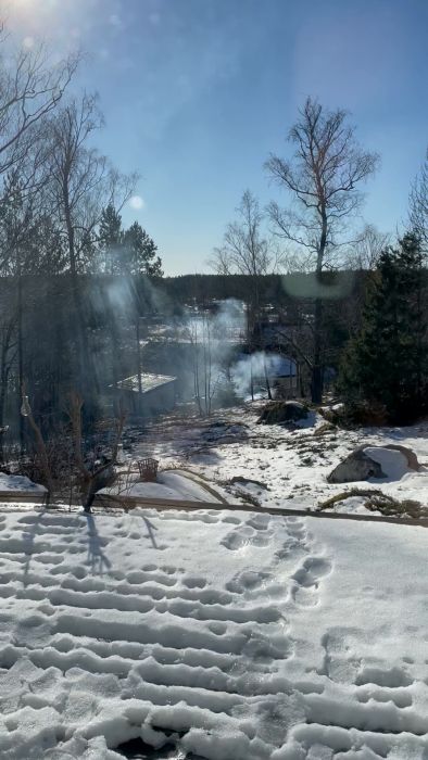 Ett snötäckt landskap med träd, solsken och smältande snö framför ett hus, sett genom ett fönster med reflektioner.