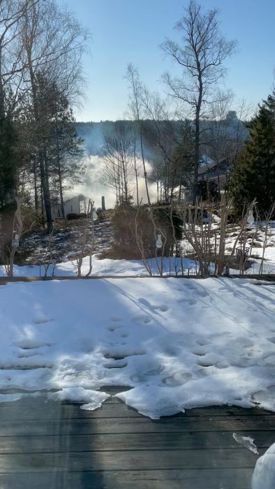 Solig vinterdag med smältande snö på en träterrass, dimma över en sjö, träd i förgrunden.
