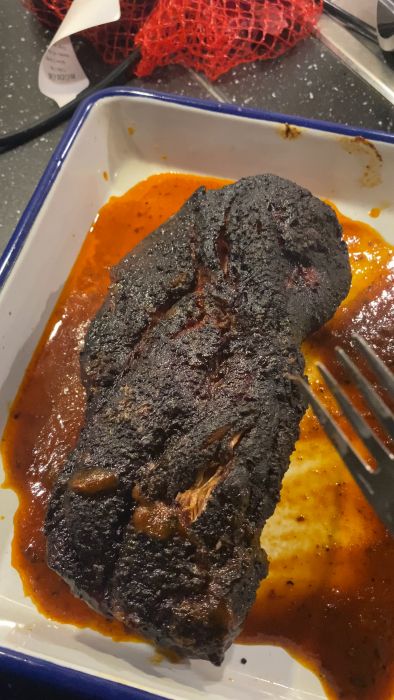 Stekt kött, möjligen överkokt, med svart yta och en gaffel, ligger i en form med sås. Köksbakgrund med packningsnät och etikett.