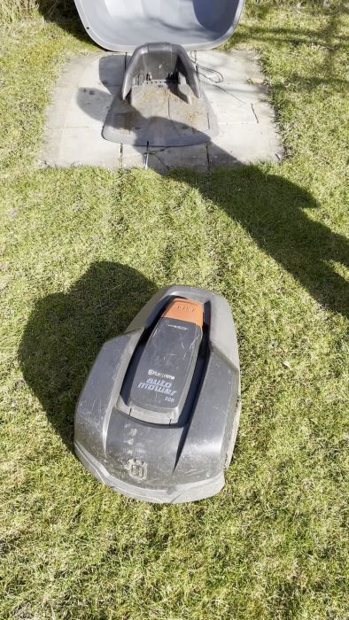 Robotgräsklippare går mot laddningsstation på betongplatta omgärdad av gräsmatta, skugga syns.