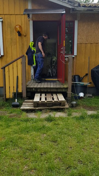 Trähus med öppen dörr, person inuti. Hund vid dörröppningen. Klädhängare, pallar och diverse föremål synliga. Kan vara bondgård eller lantligt hem.