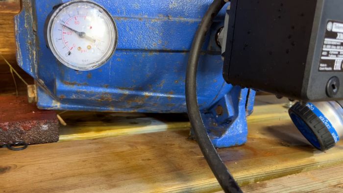 Ett blått tryckluftskompressor med mätare, svarta sladdar och golvstående på träbotten. Slitet, industriellt utseende, delvis rostig, kopplad till elektrisk utrustning.