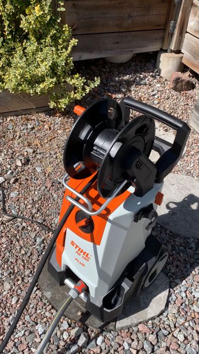 En elektrisk högtryckstvätt av märket STIHL står på en grusbelagd mark bredvid trä- och buskvegetation.
