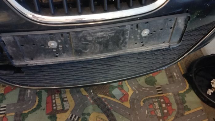 En närbild av ett bilens främre grill och registreringsskyltplats, fotograferad inomhus över ett lekmatta med vägtema. Filmen är suddig och av låg kvalitet.