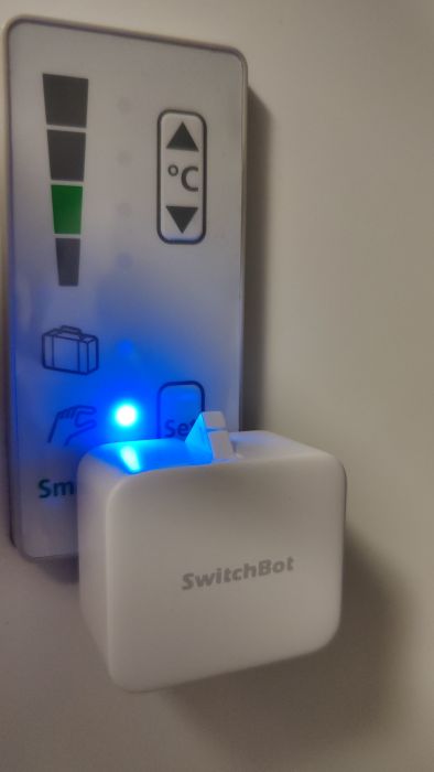 Ett vitt SwitchBot-objekt monterat på en grå yta med upplysta färgglada knappar. Verkar vara smart hem-teknik.