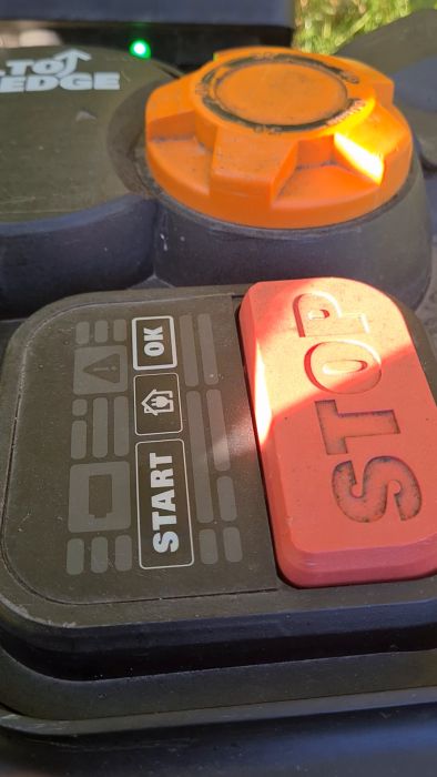 Ett maskinkontrollpanel med en orange startknapp, en röd stoppknapp och gröna ljusindikatorer i solljus.