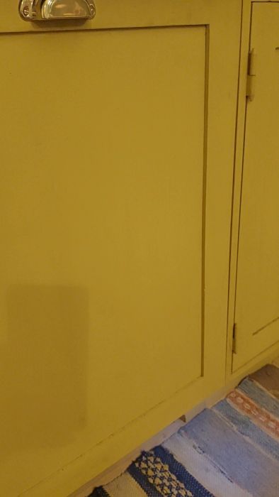 Gul skåpdörr med silverfärgat handtag, mot en gul vägg ovanför en mönstrad matta.