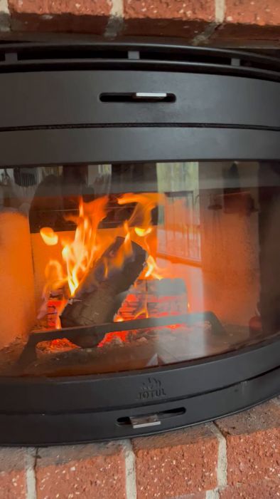 En vedeldad kamin med öppen spjällucka där elden brinner bakom ett glasfönster, omgiven av tegelstenar. Kaminen är märkt med "JØTUL".