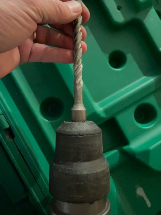 En hand håller en spiralborr ovanför ett verktygsfäste mot en grön plastbakgrund. Verktyget ser ut som ett borrfäste.